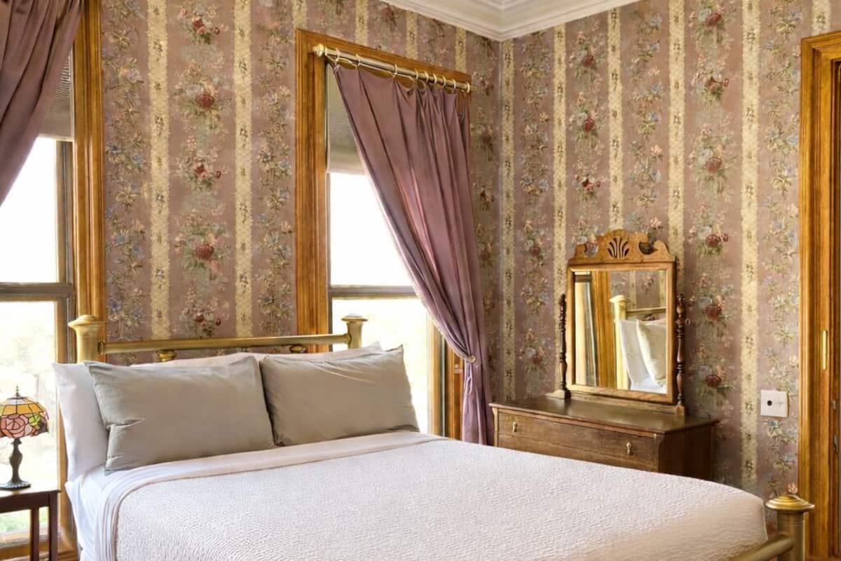 A classic four-poster bed with a wooden frame, a matching nightstand, a dresser with a mirror, and floral wallpaper.