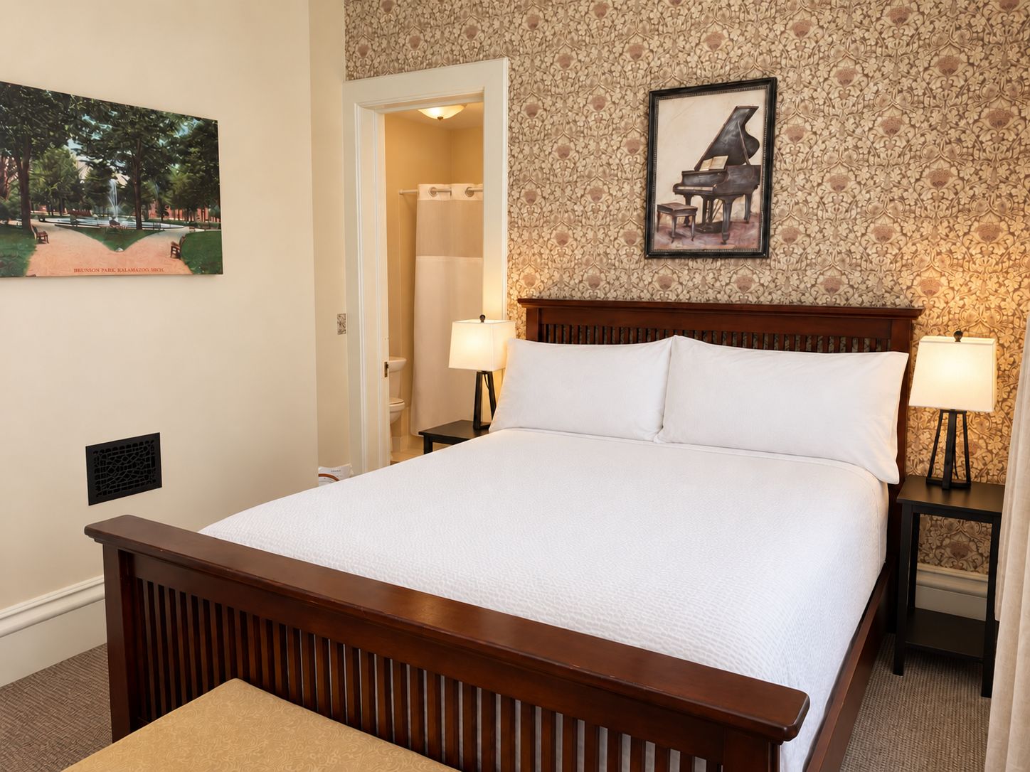 Habitación de hotel con una cama grande, dos mesitas de noche con lámparas, un cuadro enmarcado de un piano en la pared y una puerta que da a un baño.