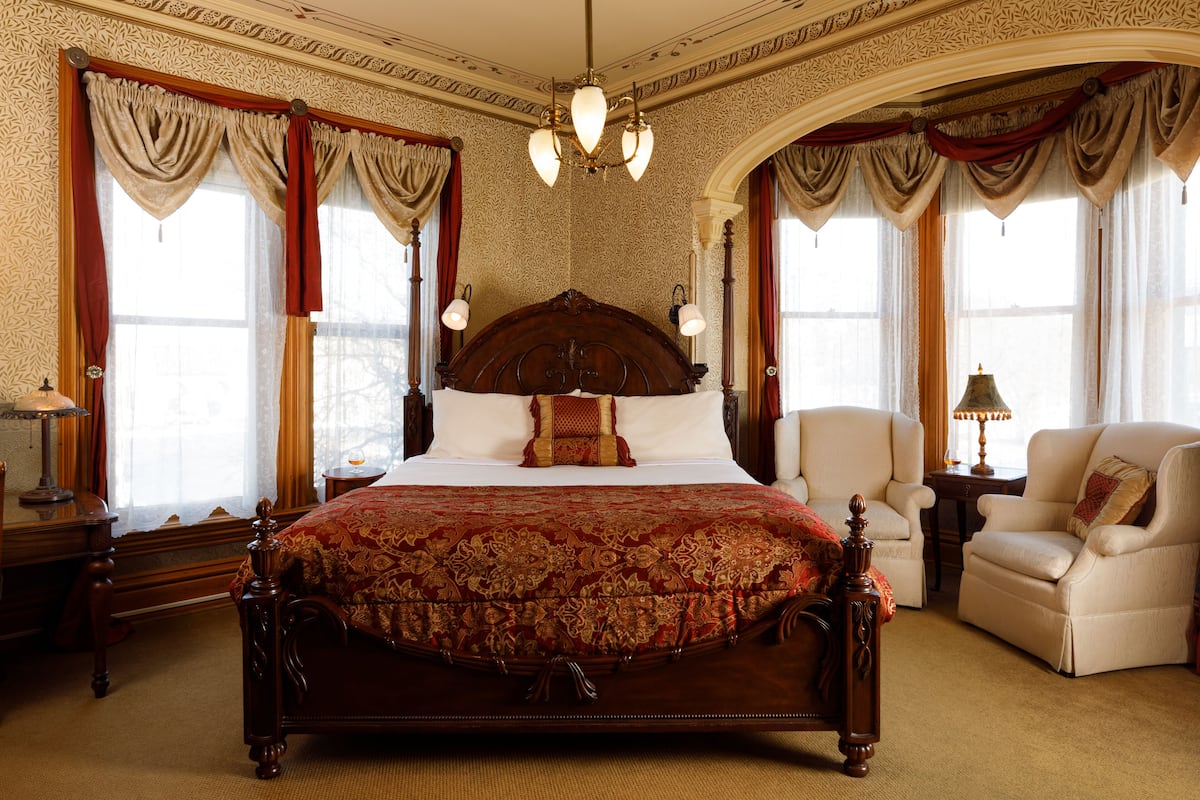 A large, ornate bed with a wooden headboard, flanked by two armchairs and a nightstand with a lamp.