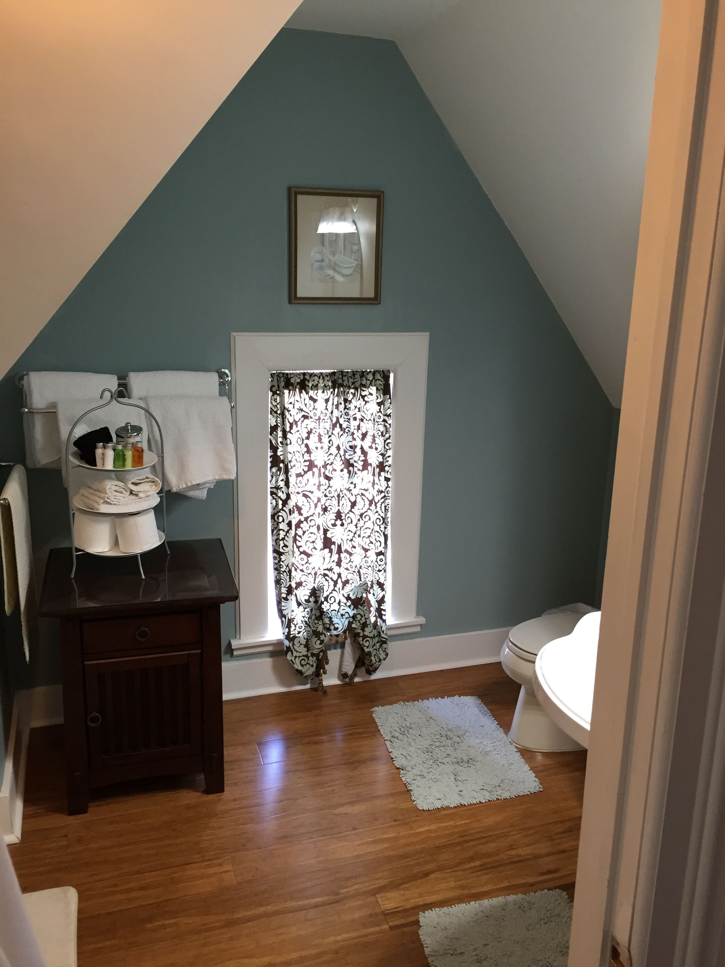 A small bathroom with a sloped ceiling, a toilet, a sink, a vanity with a mirror, and a window with patterned curtains.
