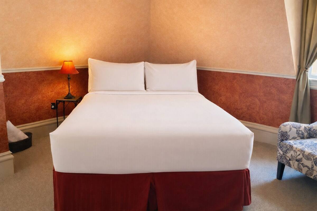 A hotel room with a large bed, a nightstand with a lamp, a chair with a floral pattern, and a window with curtains.