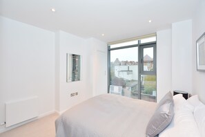 Ein modernes Schlafzimmer mit einem großen Bett, einem Fenster mit Blick auf Gebäude und einem Heizkörper an der Wand.