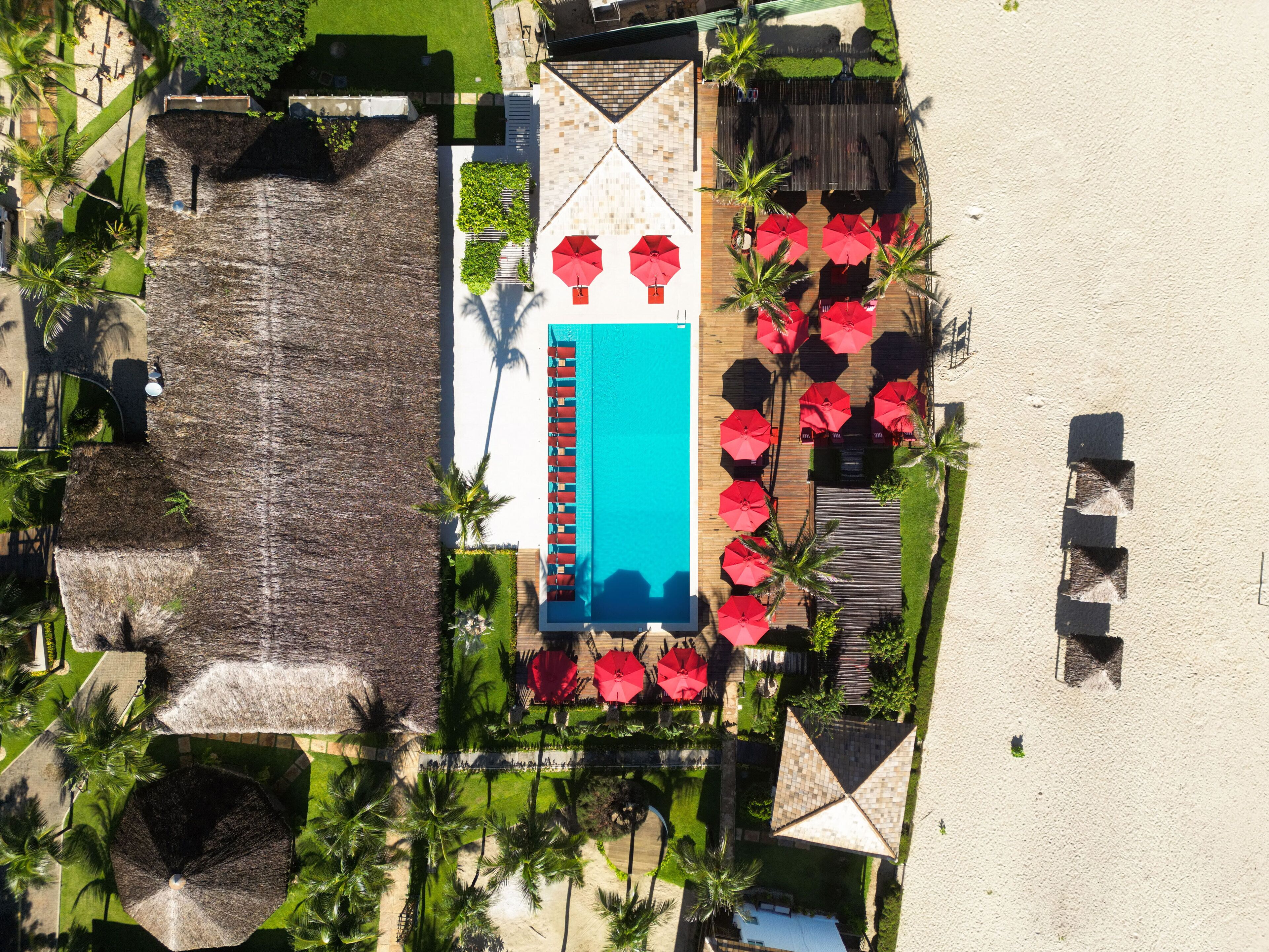 An aerial view of a resort with a swimming pool, red umbrellas, and palm trees.