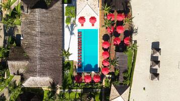 An aerial view of a resort with a swimming pool, red umbrellas, and palm trees.