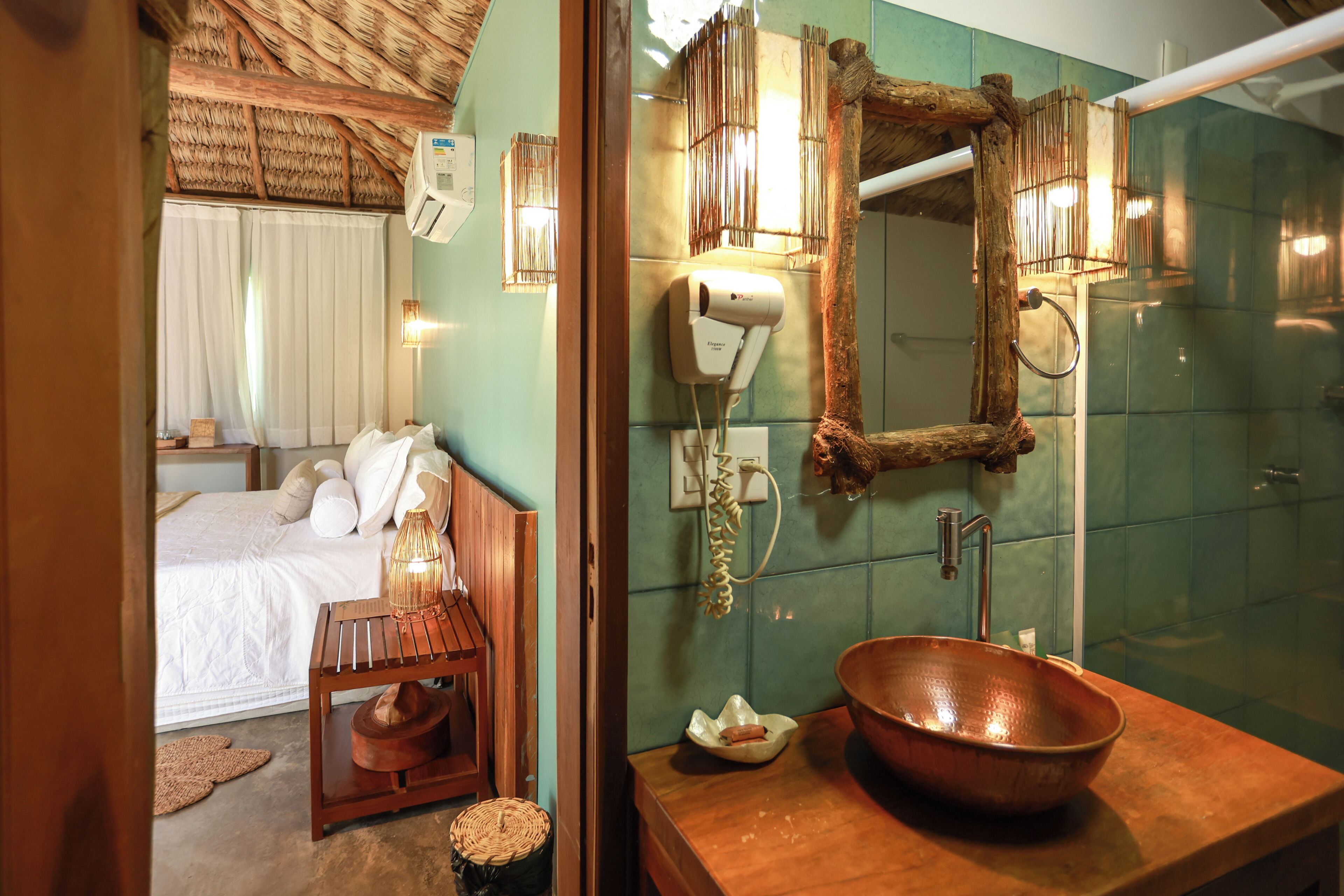 Habitación de hotel con cama, mesita de noche con lámpara, espejo y baño con lavamanos y un lavabo de cobre grande.