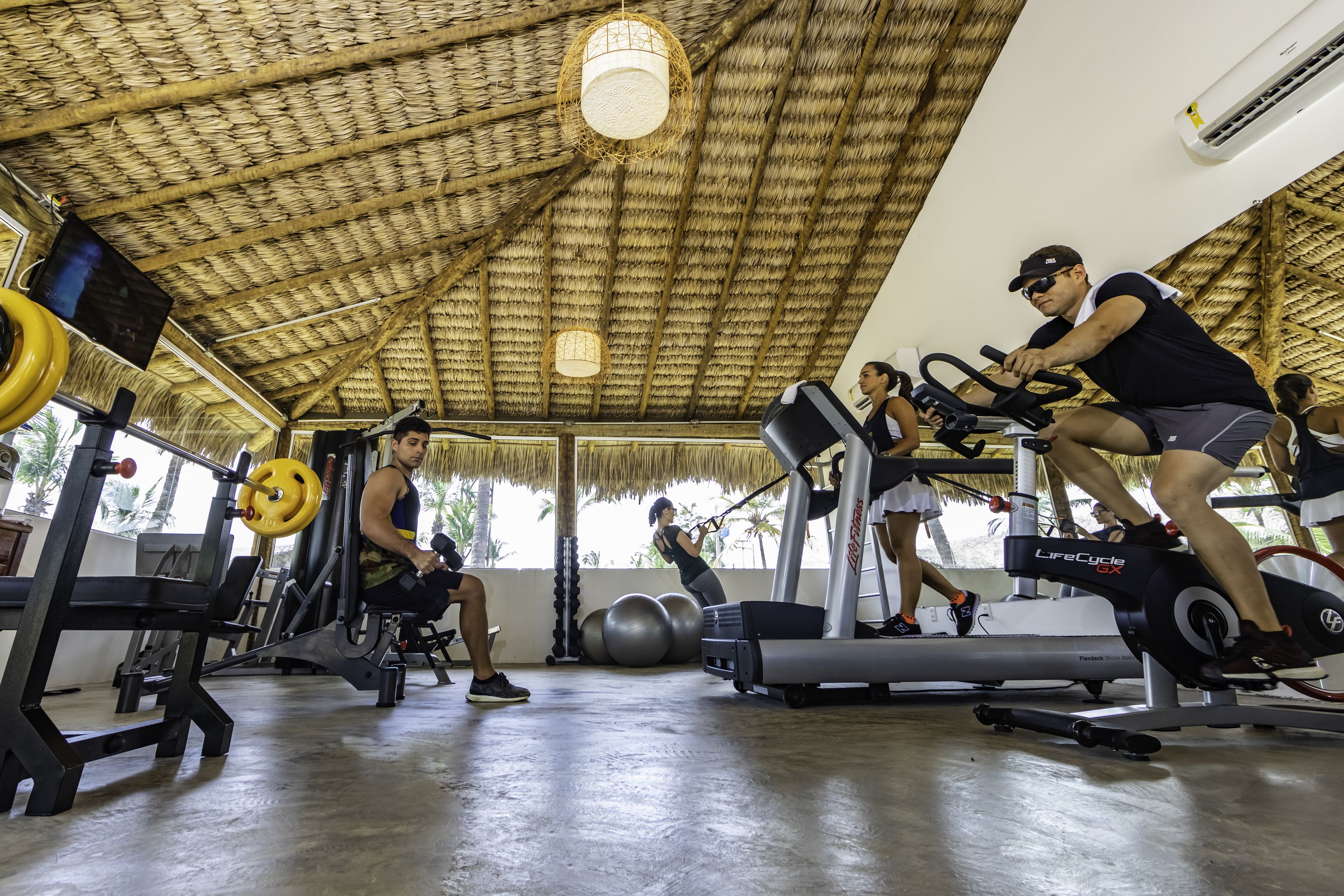 Et fitnesscenter med træningsudstyr, herunder løbebånd og styrkemaskiner, under et stråtækt tag.