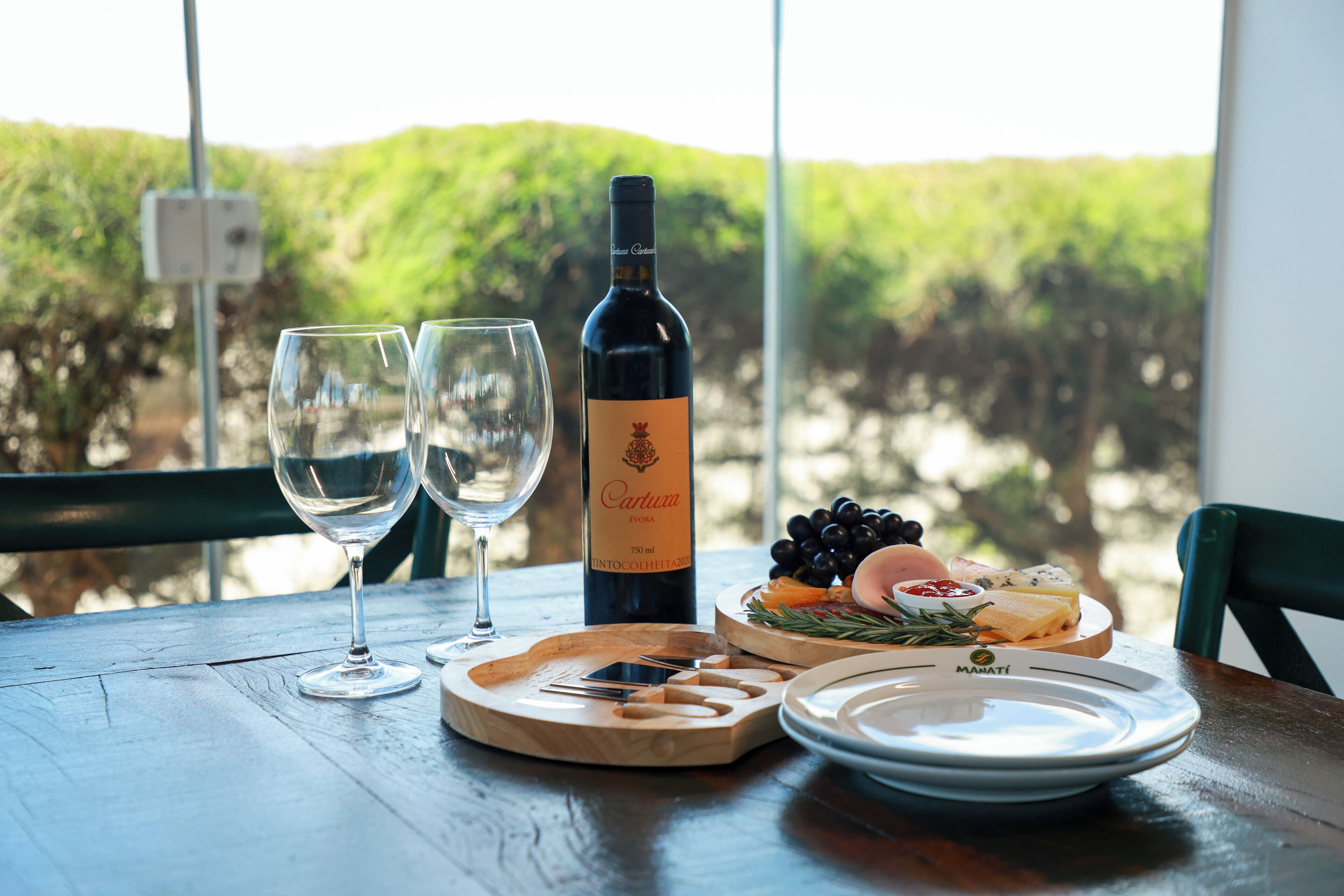 Une table avec une bouteille de vin, deux verres, une planche en bois avec du fromage et des raisins, et une assiette avec un couteau et une fourchette.