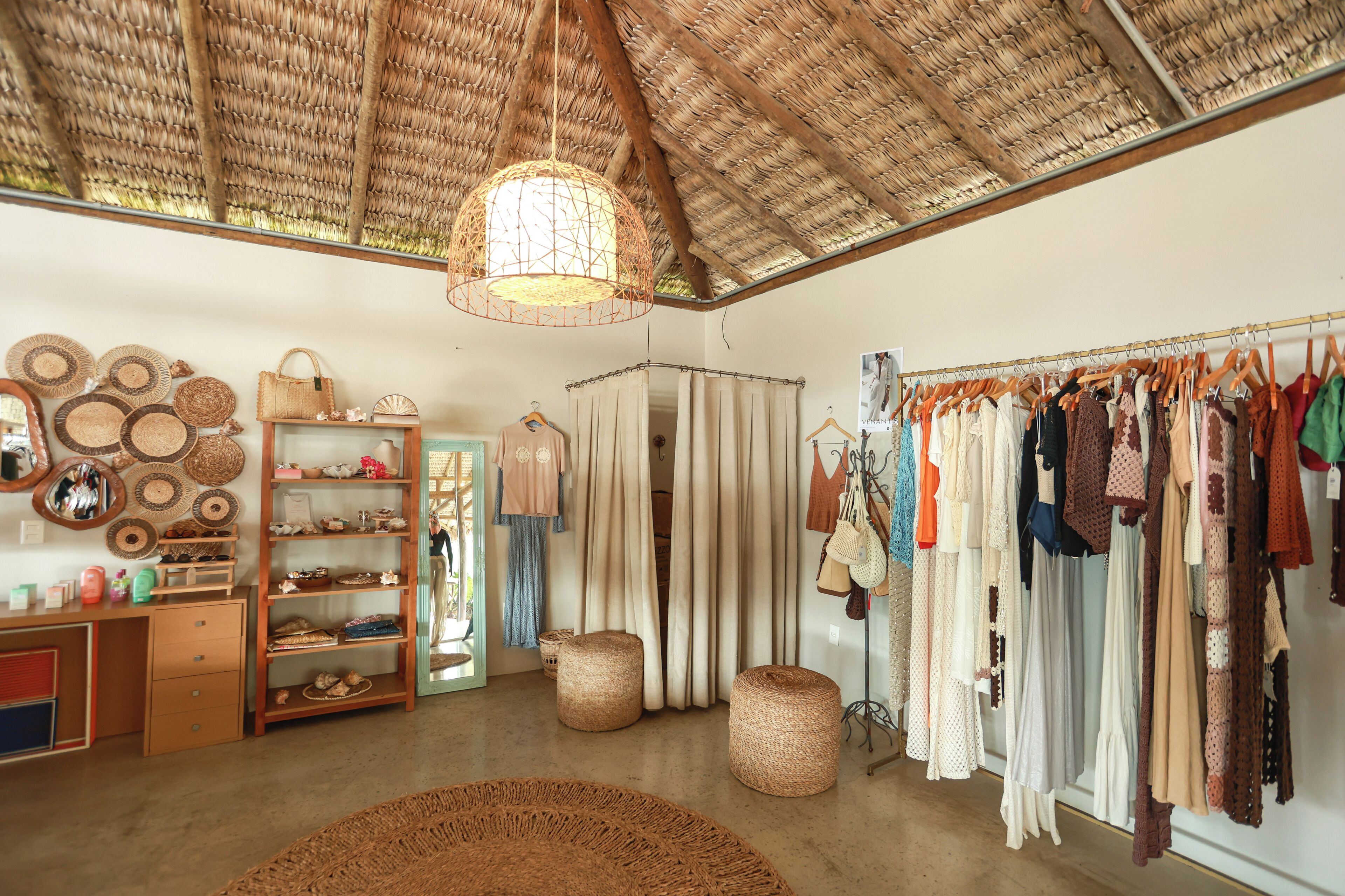 A boutique with a rustic interior, featuring a clothing rack, shelves with various items, and a woven ceiling.