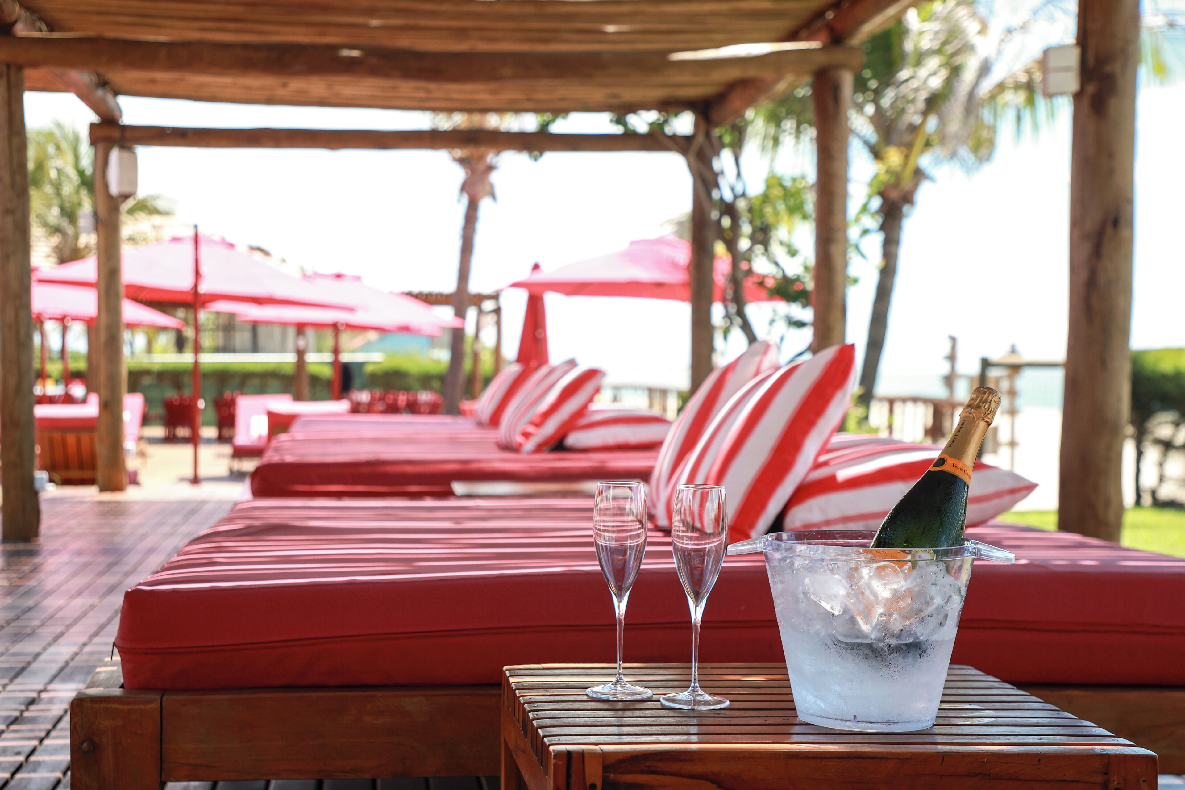 A wooden table with a bottle of champagne, two glasses, and a glass of water on a red cushioned bench.