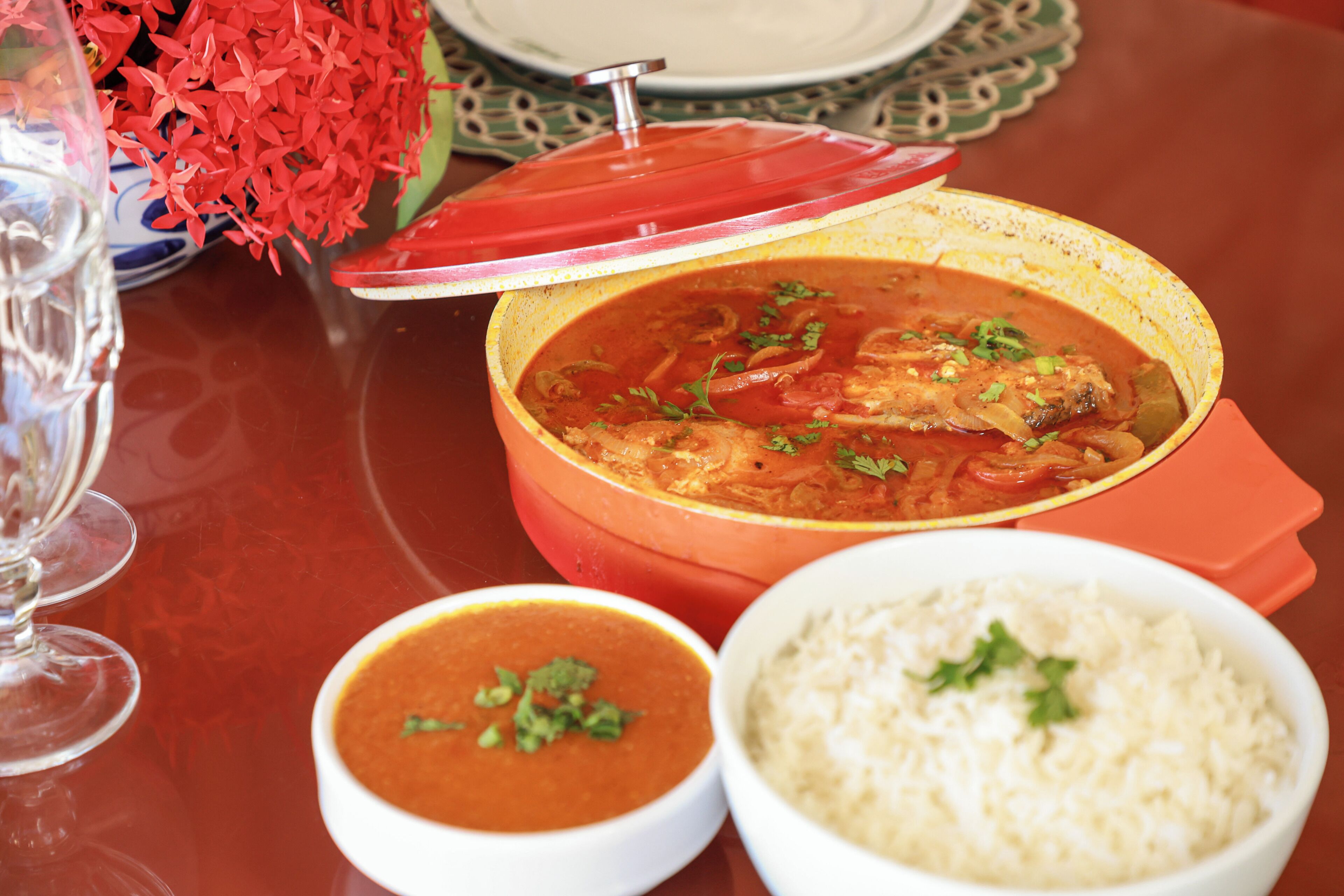 A table with a red tablecloth, a bowl of rice, a bowl of soup, and a pot of curry.