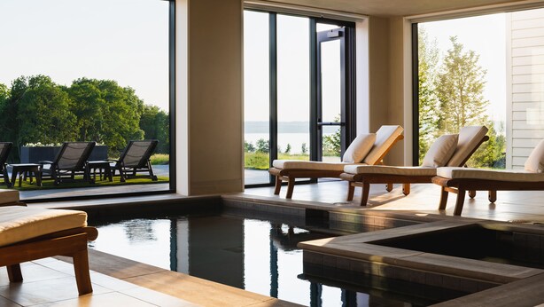 Un espace piscine intérieur moderne, doté de chaises longues et offrant une vue sur l’extérieur.