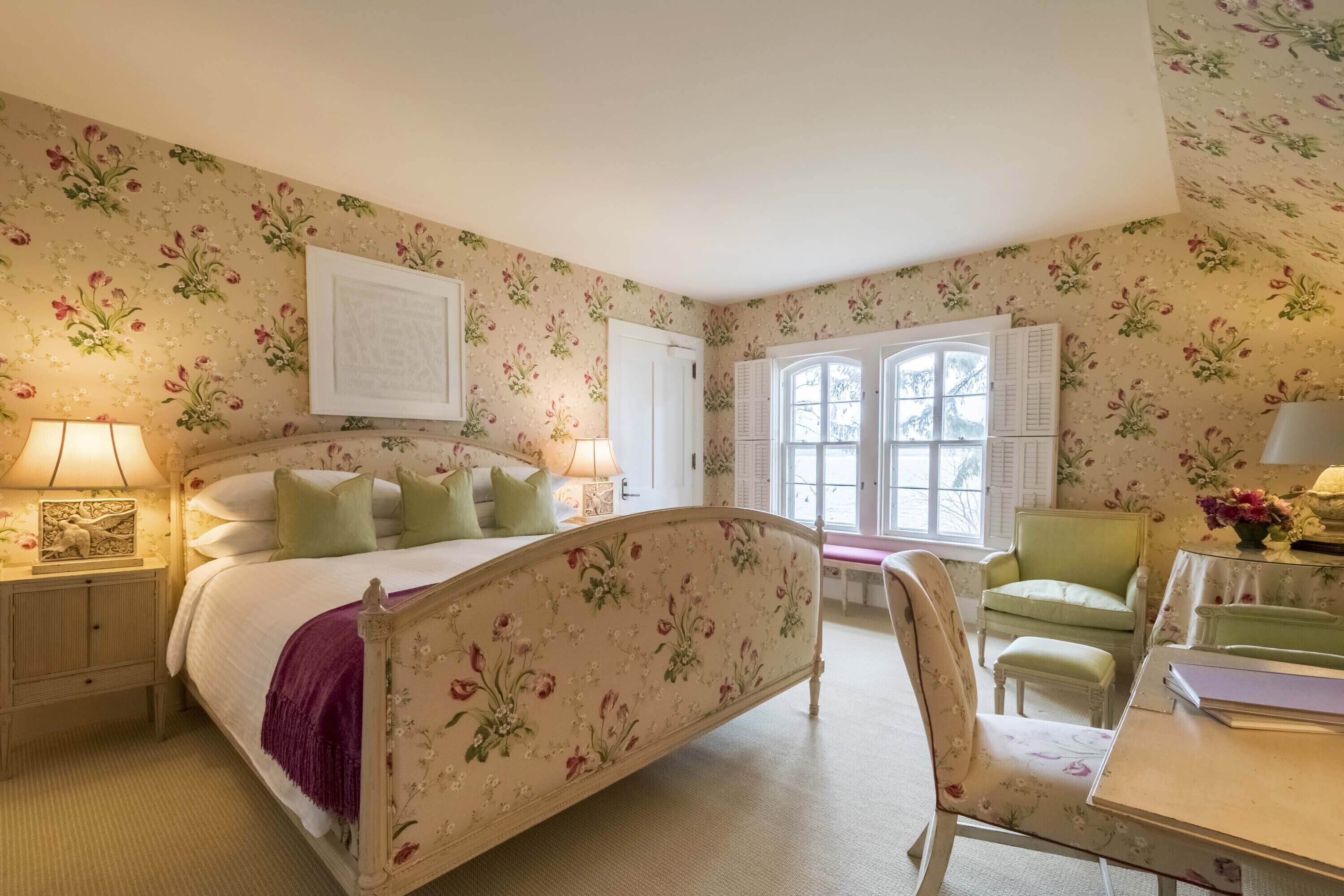 Un dormitorio con papel tapiz floral, una cama con cabecera, una mesita de noche, una silla y una ventana con contraventanas.