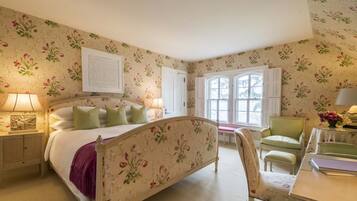Un dormitorio con papel tapiz floral, una cama con cabecera, una mesita de noche, una silla y una ventana con contraventanas.