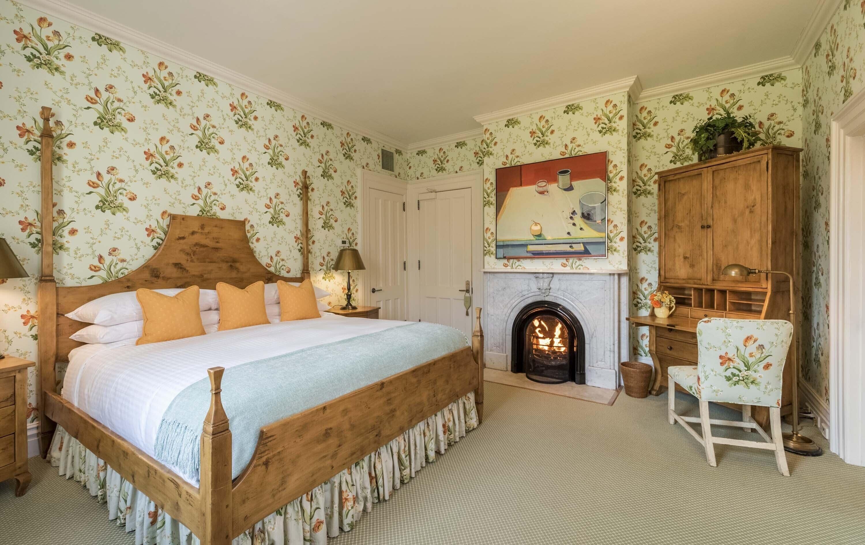 A bedroom with a wooden four-poster bed, a fireplace, floral wallpaper, and a wooden cabinet.