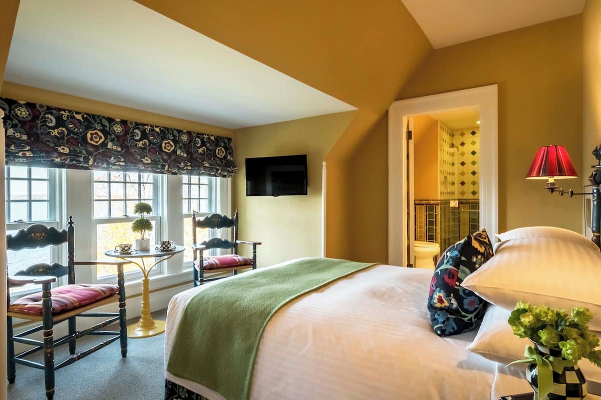 A hotel room with a bed, a TV mounted on the wall, a small table with chairs, and a window with a view.