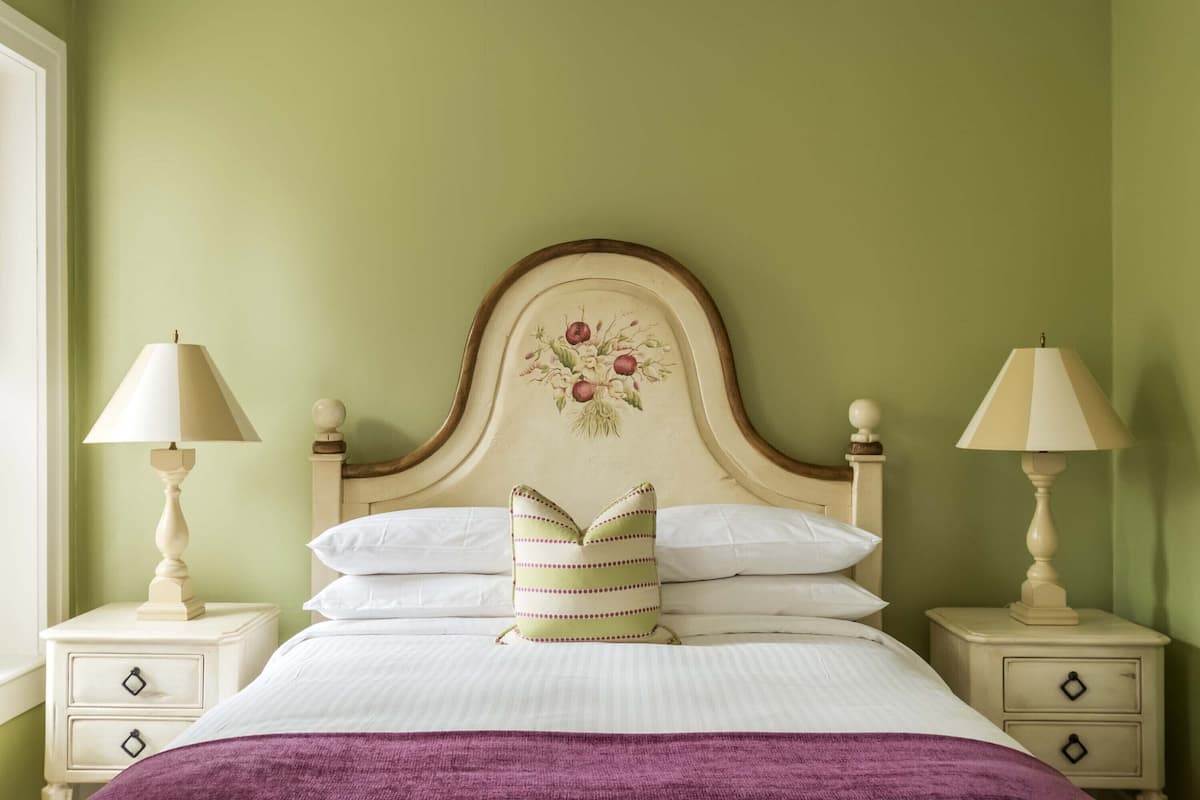 A neatly made bed with white linens and a purple throw, flanked by two bedside tables with lamps, set against a green wall.