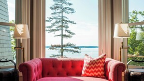 Un canapé rouge orné d’un coussin à motifs chevrons, placé devant une fenêtre donnant sur un arbre et un plan d’eau.