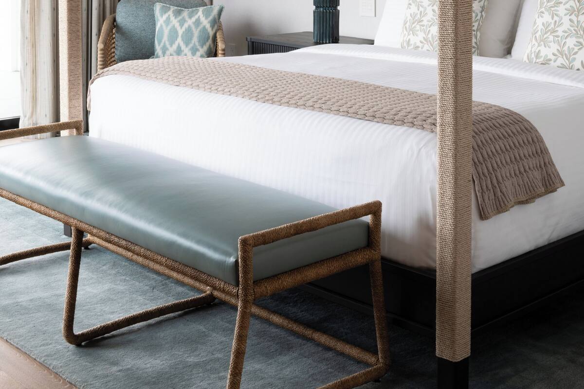 A four-poster bed with a bench, a bedside table with a lamp, and a framed artwork on the wall.