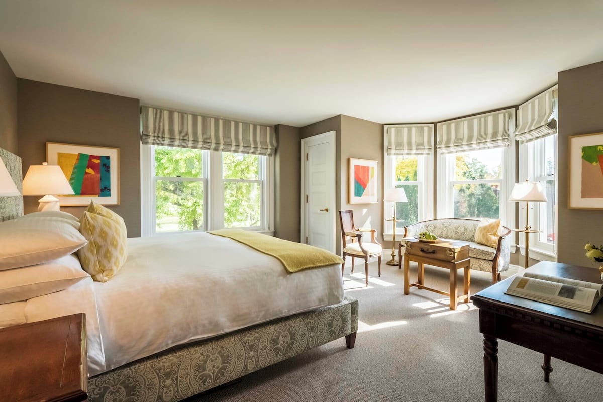 A bedroom with a large bed, a desk, and a chair. There are windows with curtains, a door, and artwork on the walls.