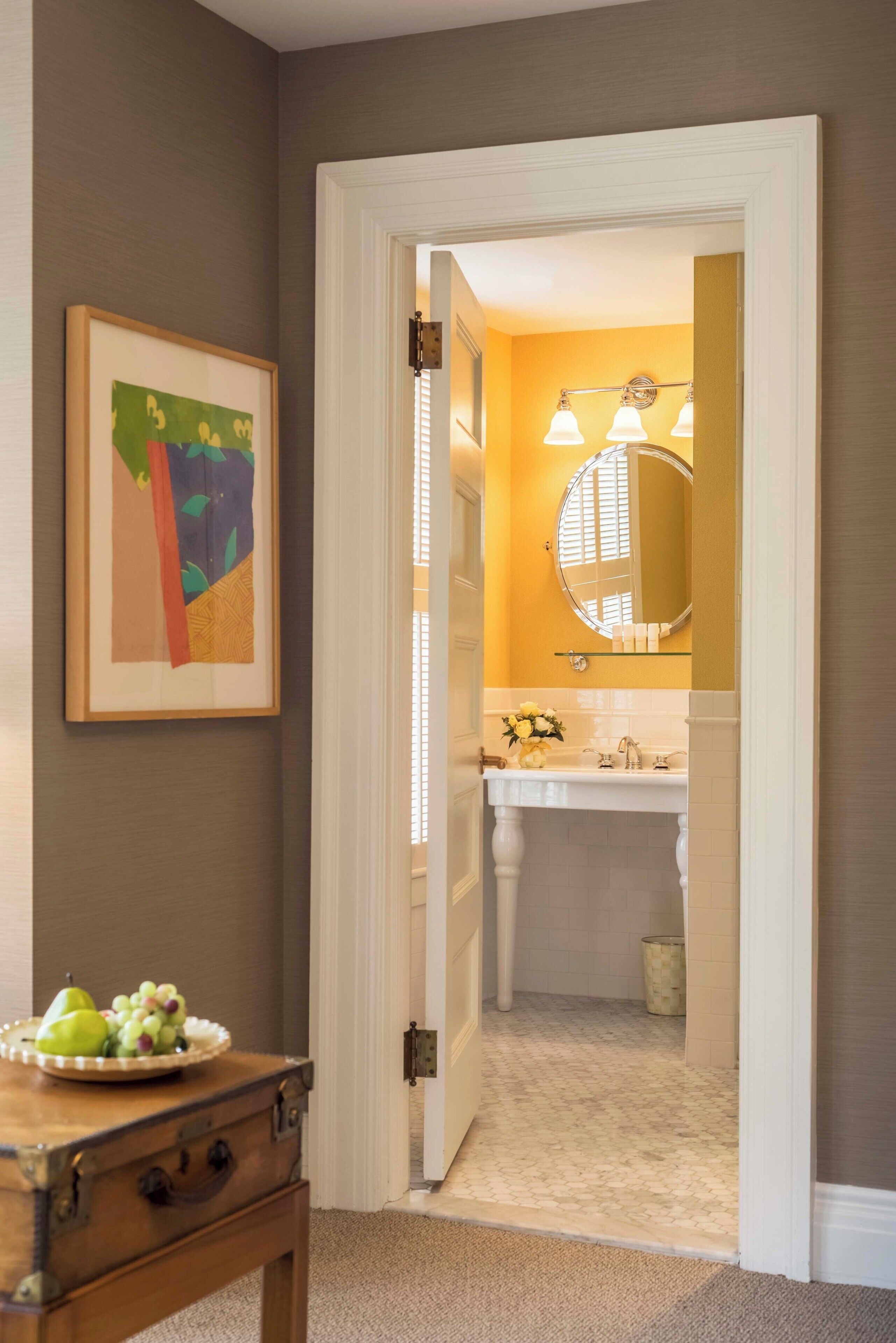 A hallway leading to a bathroom with a white sink, a round mirror, and a framed abstract painting on the wall.