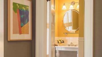 A hallway leading to a bathroom with a white sink, a round mirror, and a framed abstract painting on the wall.