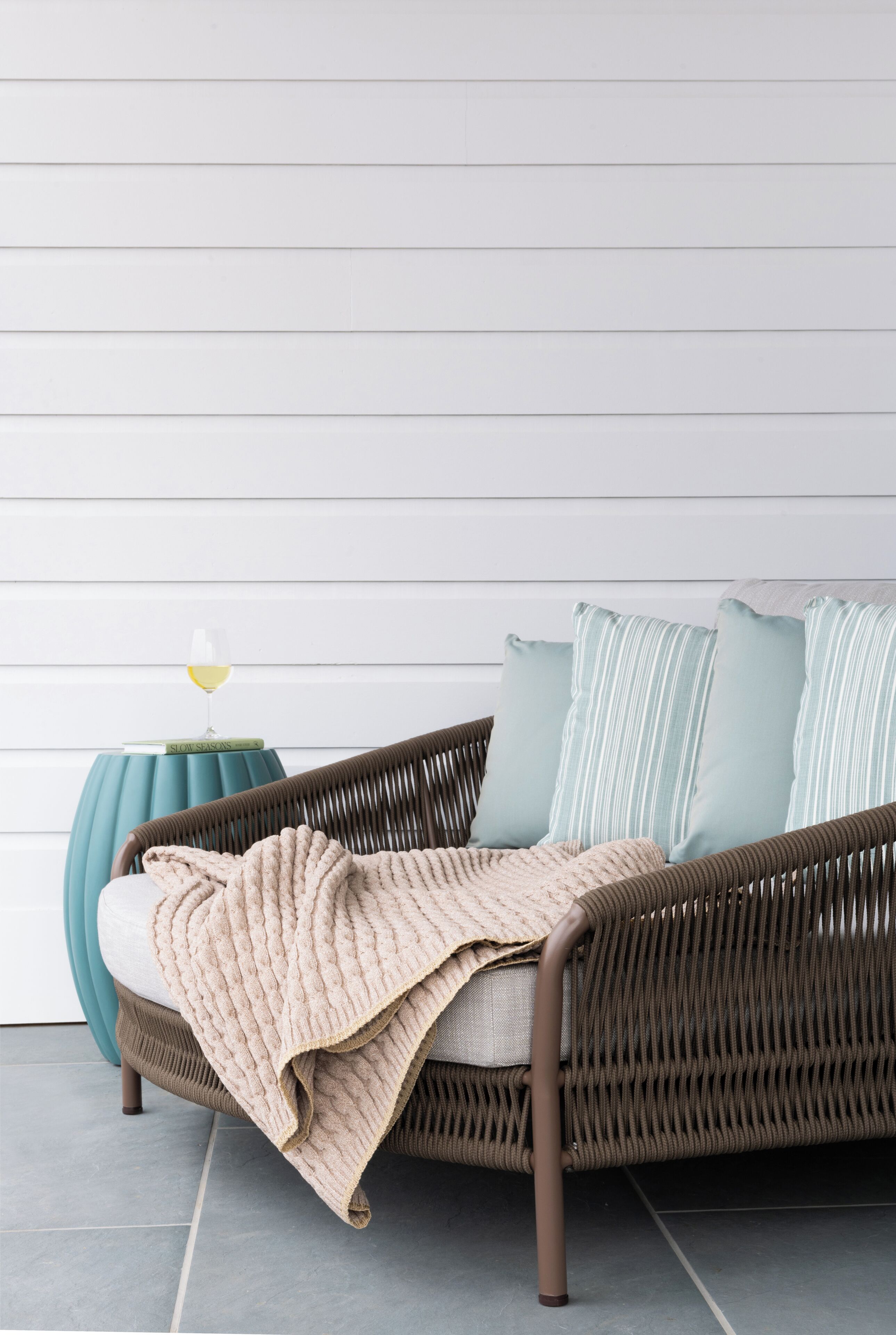 A wicker sofa with a beige blanket, light blue pillows, and a glass of wine on a side table.