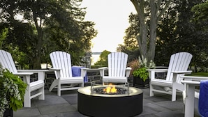 Un foyer extérieur circulaire, entouré de chaises Adirondack blanches, est installé sur une terrasse en pierre offrant une vue sur un lac et des arbres.