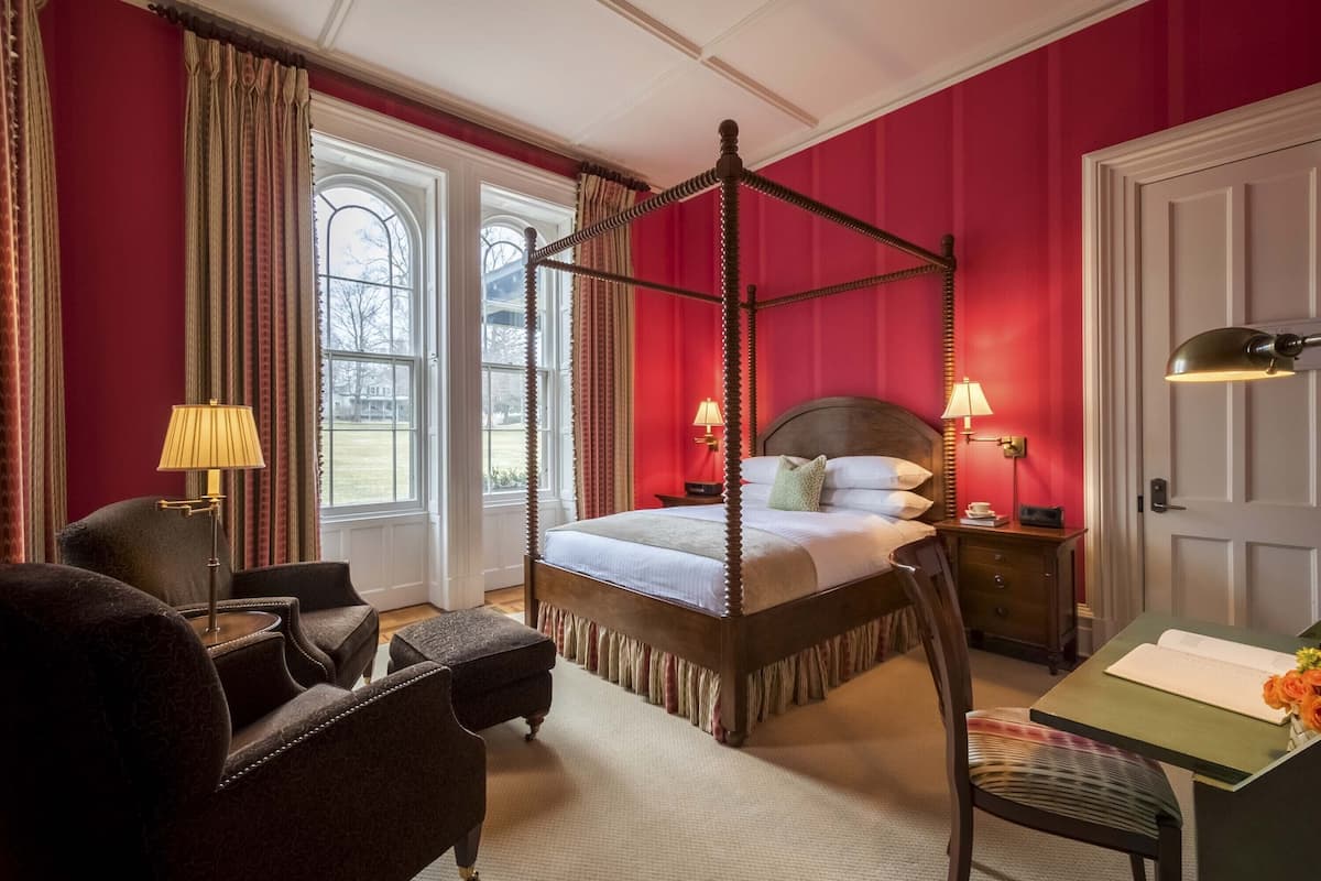 A four-poster bed with a wooden frame, a green desk with a book and flowers, two lamps, a chair, and a window with curtains.