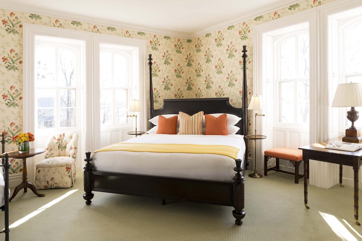 A four-poster bed with a yellow throw, a chair with floral upholstery, and a desk with a lamp.