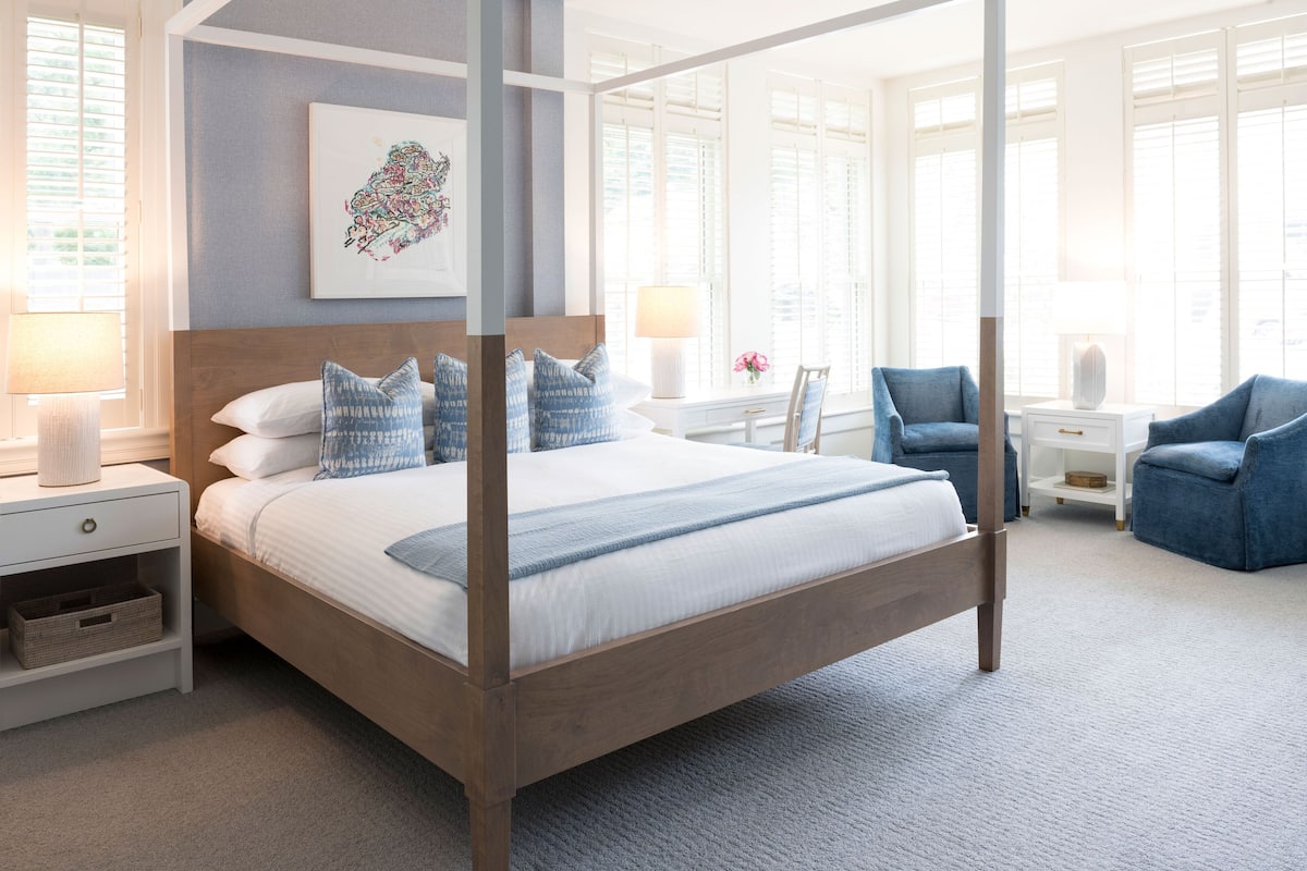A four-poster bed with a wooden frame, white bedding, and blue pillows. There is a white nightstand with a lamp and a small basket. A blue armchair is visible in the background.