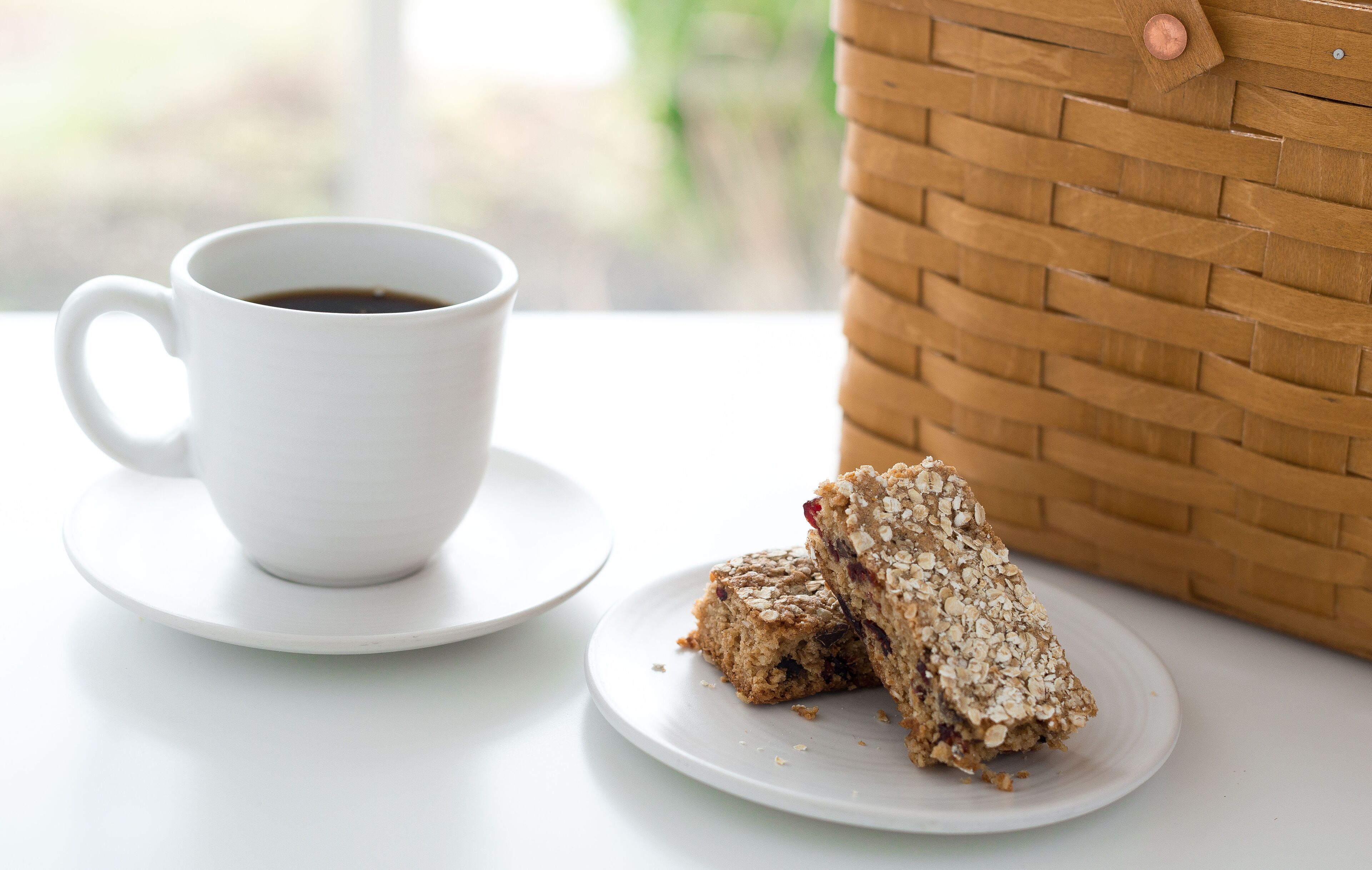 Una taza de café blanca sobre un plato, un plato con dos porciones de granola y una cesta tejida sobre una mesa.