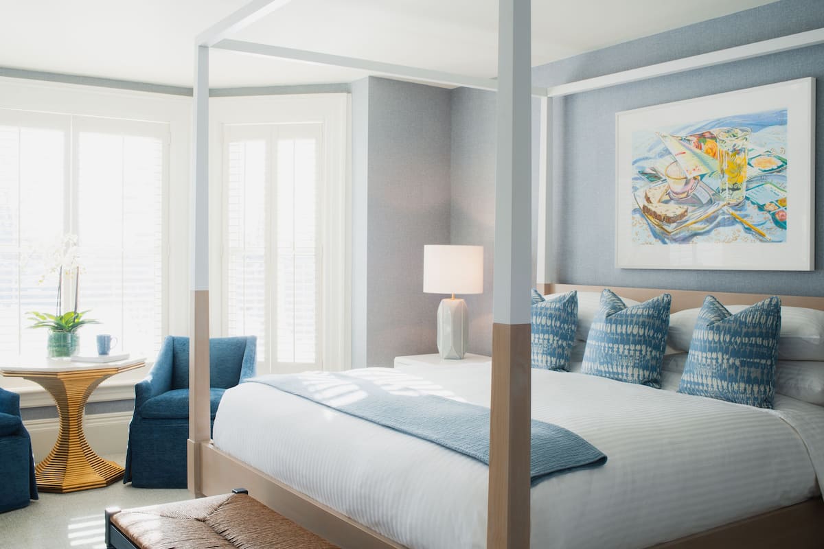 A modern hotel room with a large bed, a bedside table, a chair, and a window with blinds.