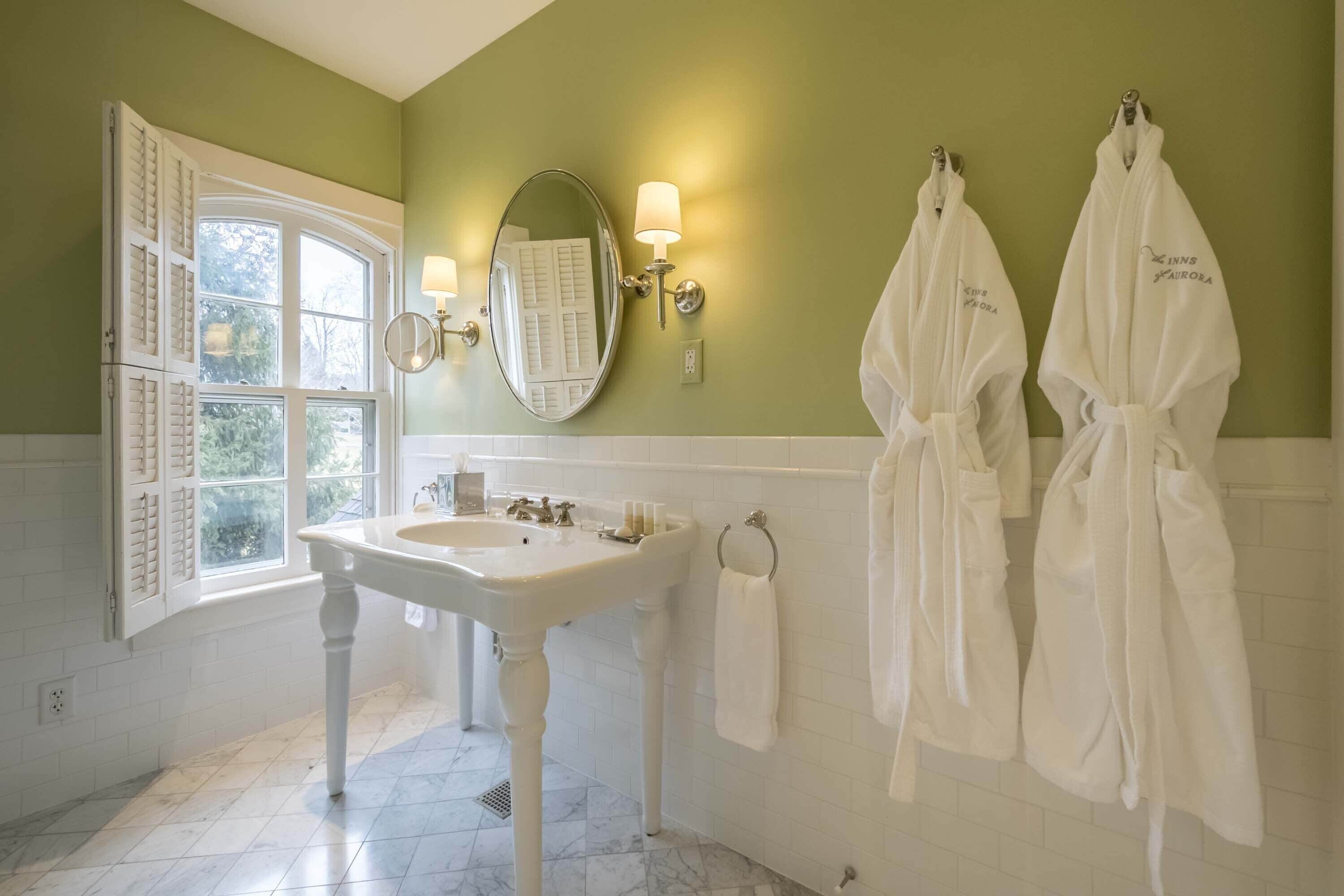 A bathroom with a white sink, two white robes hanging on the wall, and a window with shutters.