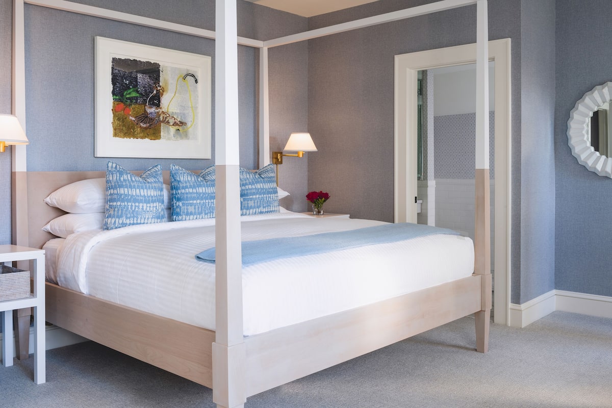 A neatly arranged bedroom with a four-poster bed, a bedside table with a lamp, and a framed artwork on the wall.