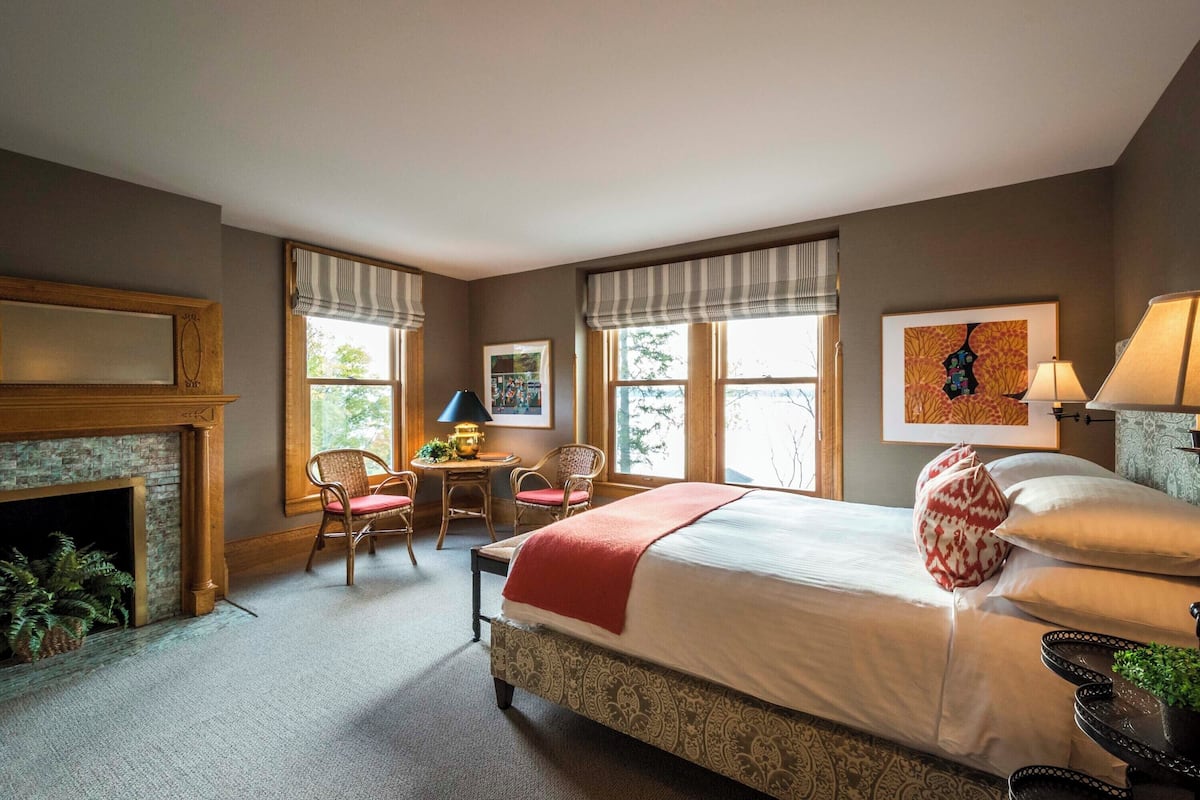 A bedroom with a large bed, a fireplace, a desk with chairs, and large windows.