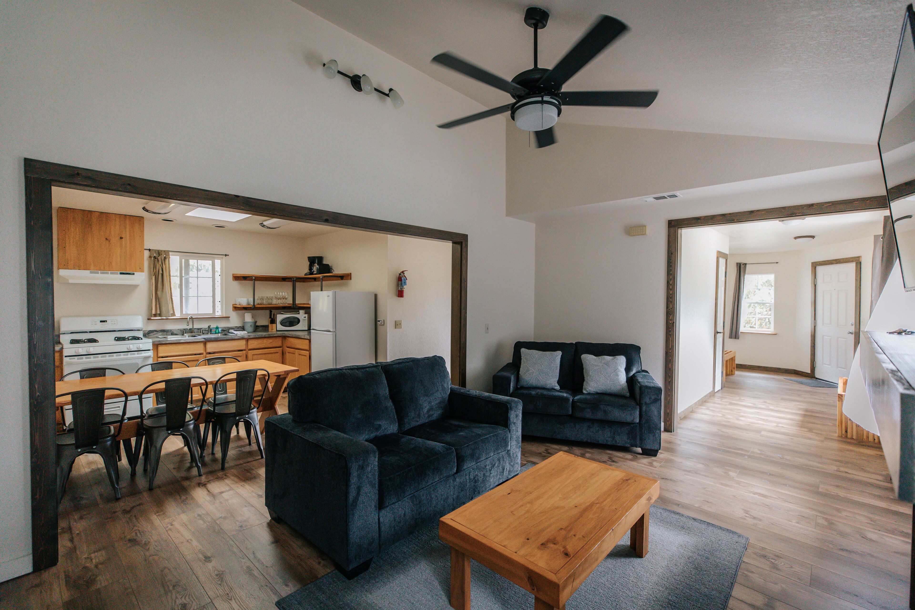 Ein geräumiger Wohnbereich mit einem dunkelblauen Sofa, einem Holztisch und einem Essbereich mit Tisch und Stühlen. Ein Deckenventilator ist sichtbar, und der Boden ist mit Holzdielen belegt.