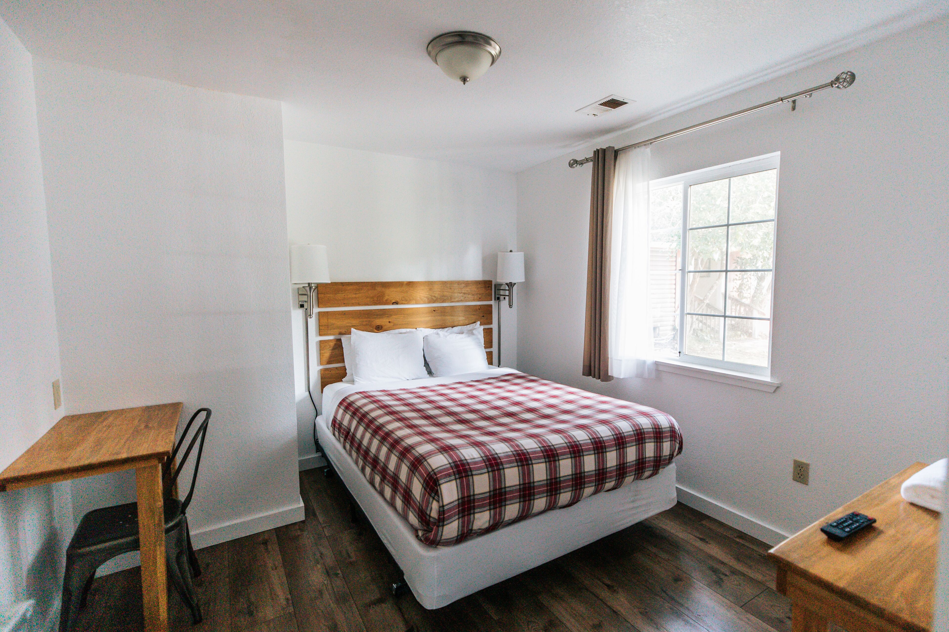 Un dormitorio con cabecera de madera, cama individual con colcha a cuadros, mesita de noche con lámpara y ventana con cortinas.