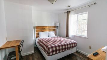 Un dormitorio con cabecera de madera, cama individual con colcha a cuadros, mesita de noche con lámpara y ventana con cortinas.