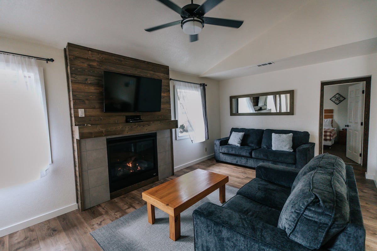 A living room with a fireplace, a television mounted on a wooden wall, a sofa, and a ceiling fan.