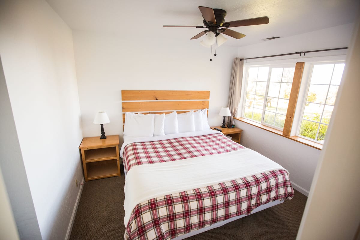 A hotel room with a wooden headboard, a bed with white and red plaid bedding, a nightstand with a lamp, a window with a view of greenery, and a ceiling fan.