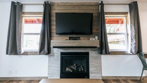 Una sala de estar con televisor de pantalla plana sobre la chimenea, paredes de madera y dos ventanas con cortinas.