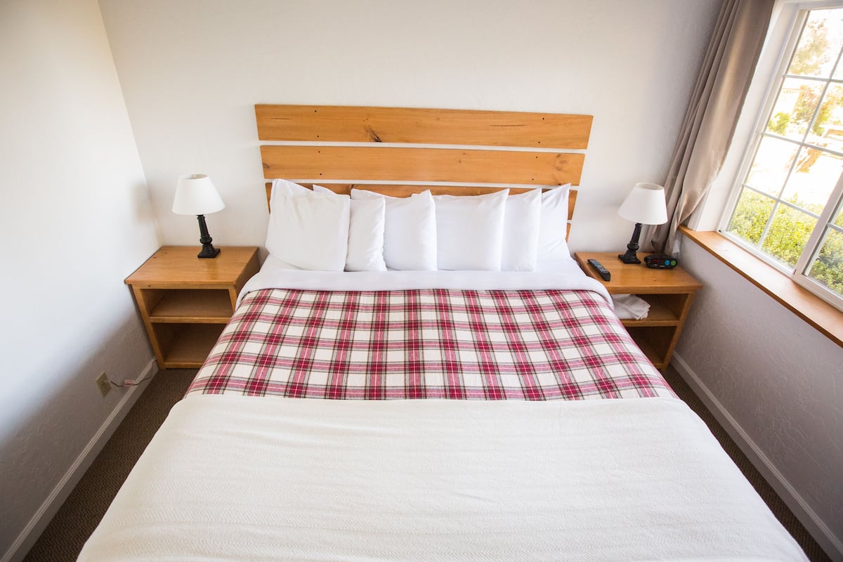 A neatly made bed with white bedding and a plaid skirt, wooden headboard, bedside tables with lamps, and a window with a view.