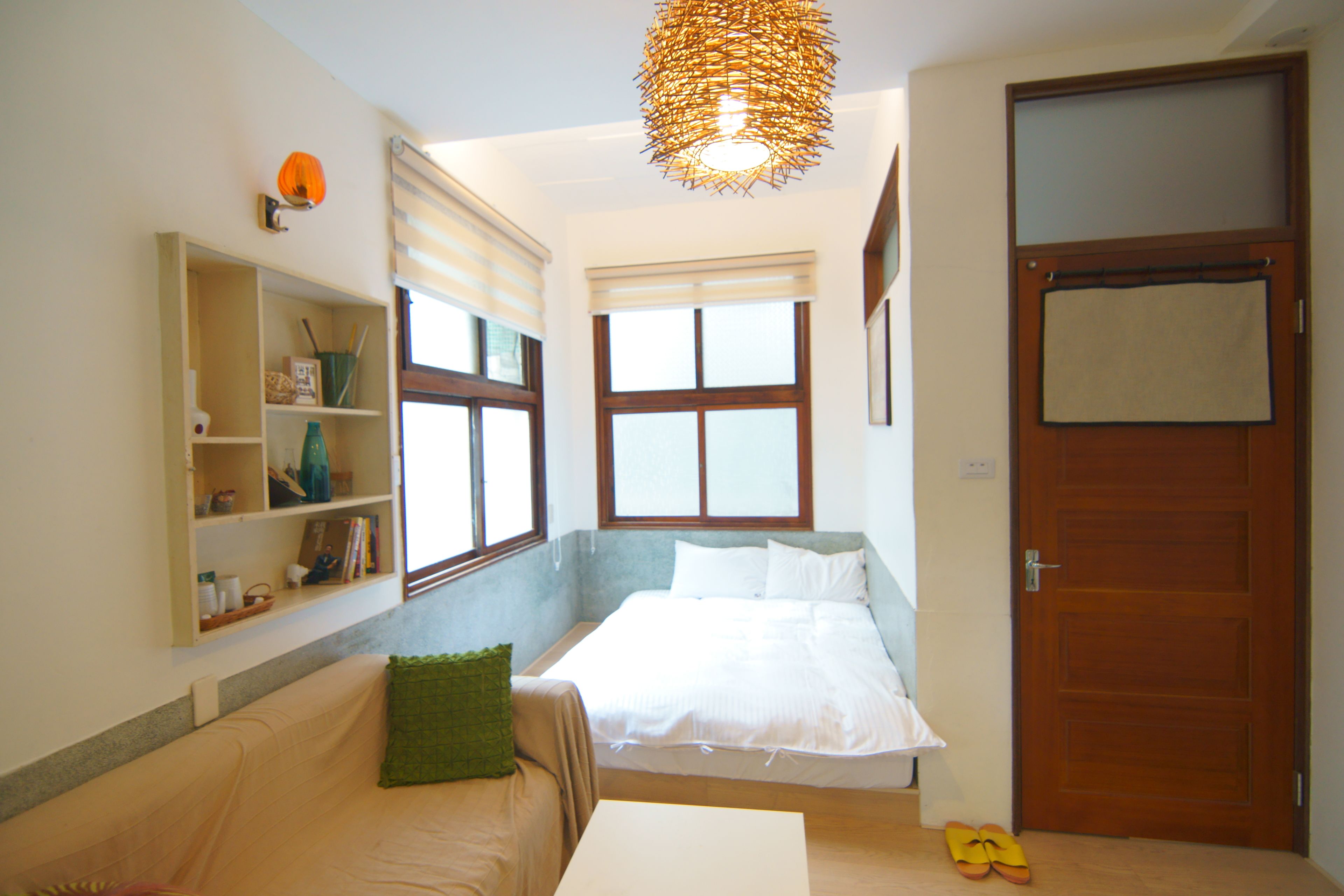 A bedroom with a bed, a bedside table, a shelf with books and a lamp, and a door.