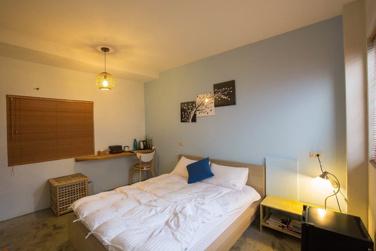 A bedroom with a bed, bedside table, lamp, wall art, and a window with blinds.