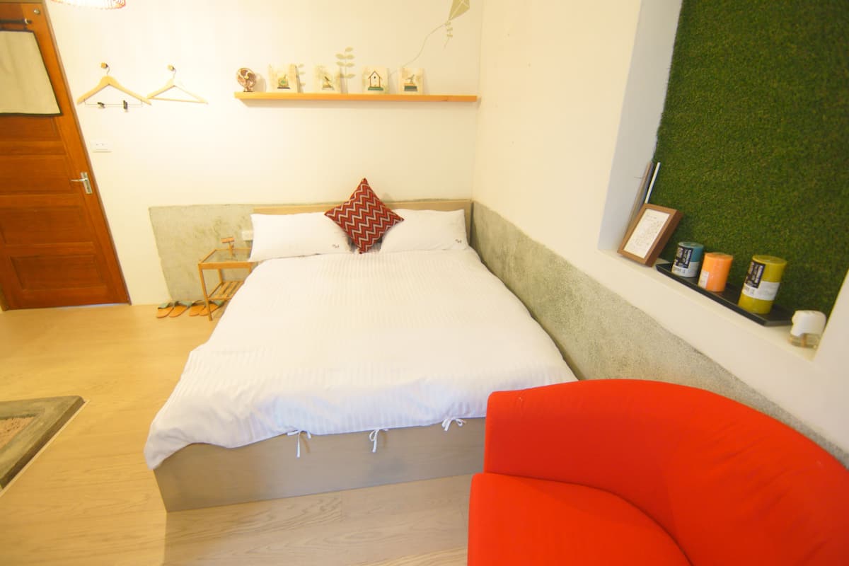 A bedroom with a bed, a red chair, a wooden door, and a shelf with small items.