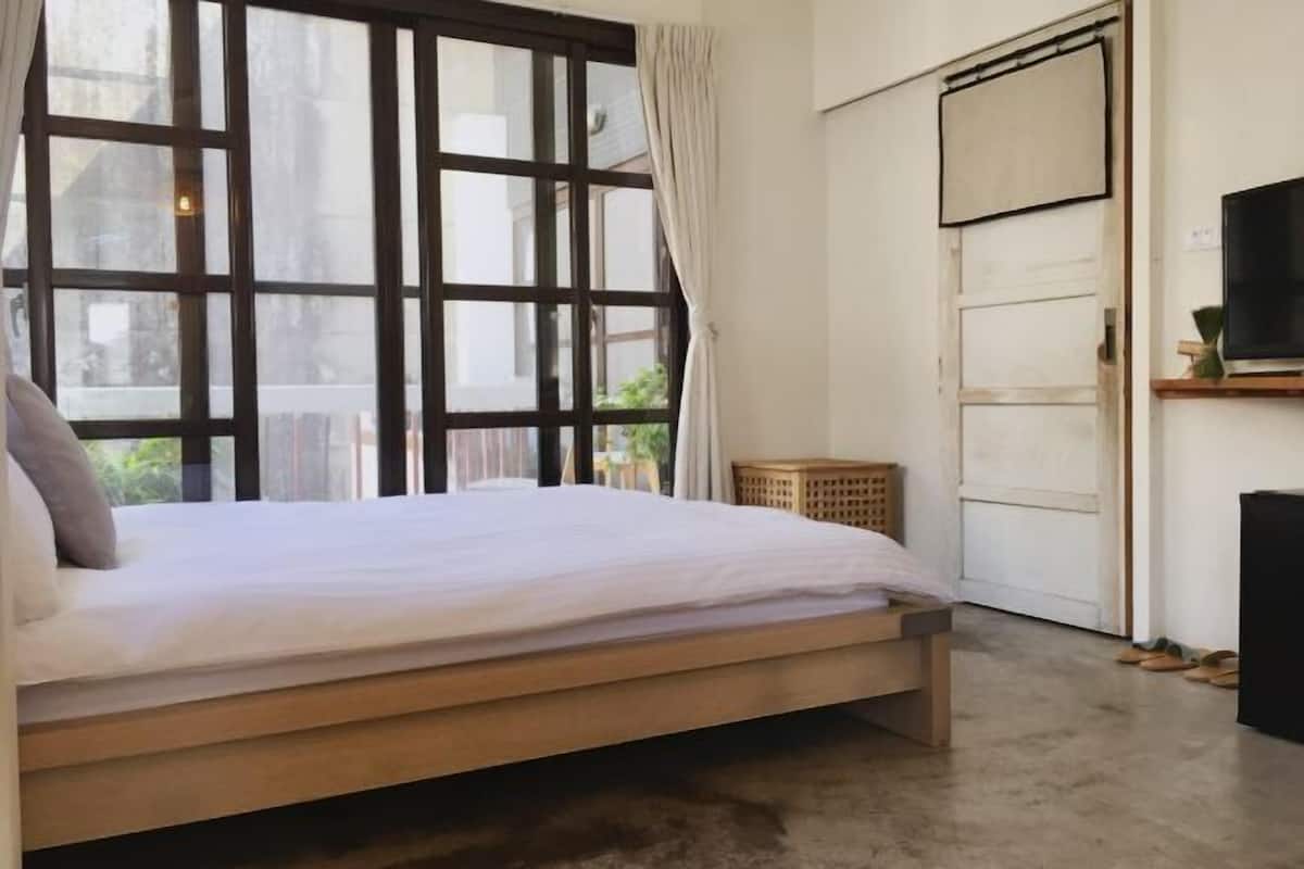 A bedroom with a wooden bed, a large window with curtains, a TV on a stand, and a door with a view of the outside.