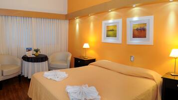 A hotel room with a bed, two chairs, a small table with a vase and a sign, two bedside lamps, and two framed pictures on the wall.