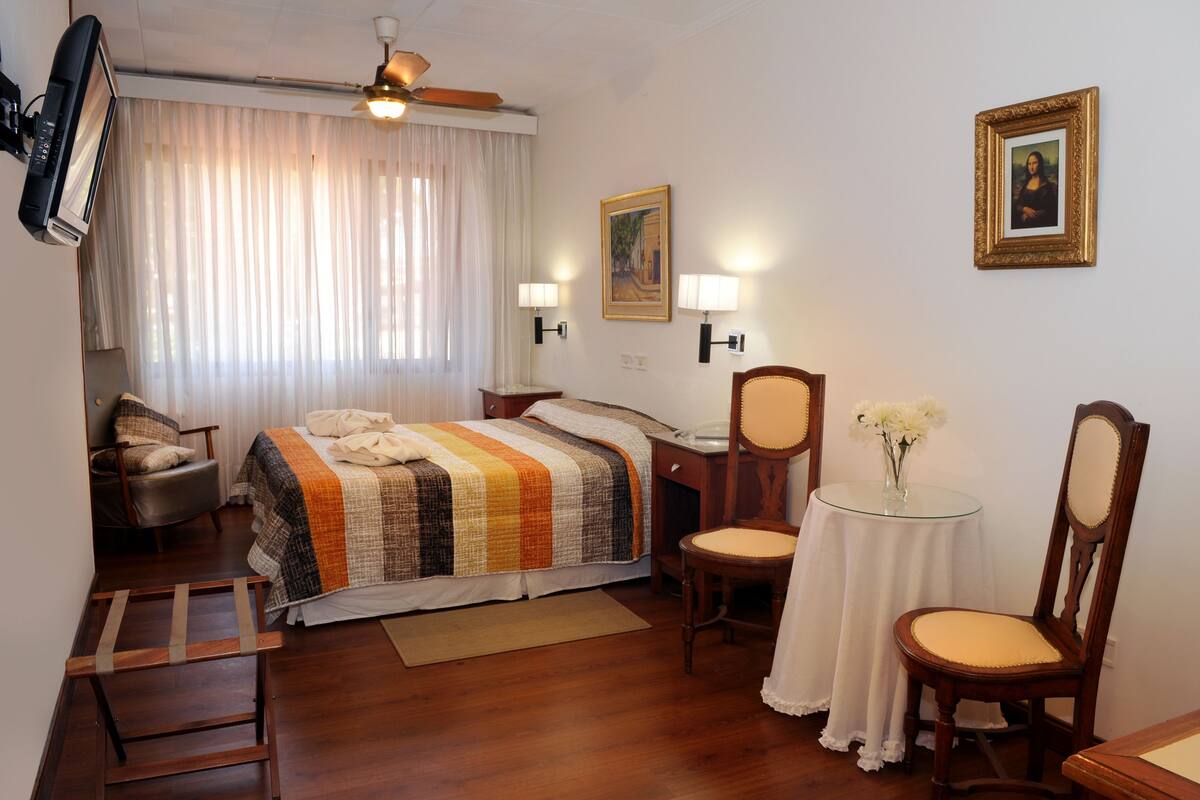A hotel room with a bed, a chair, a desk, a ceiling fan, and a painting on the wall.