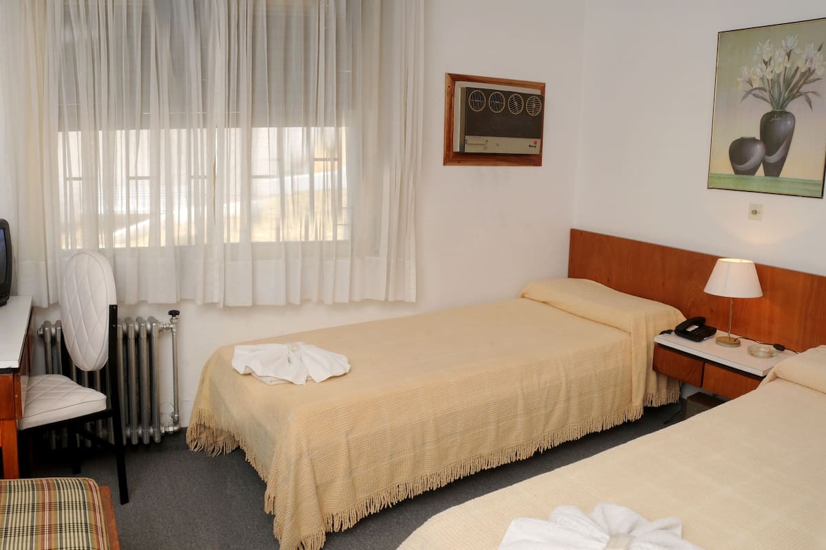 A hotel room with a bed, a desk with a television, a chair, and a bedside table with a lamp.
