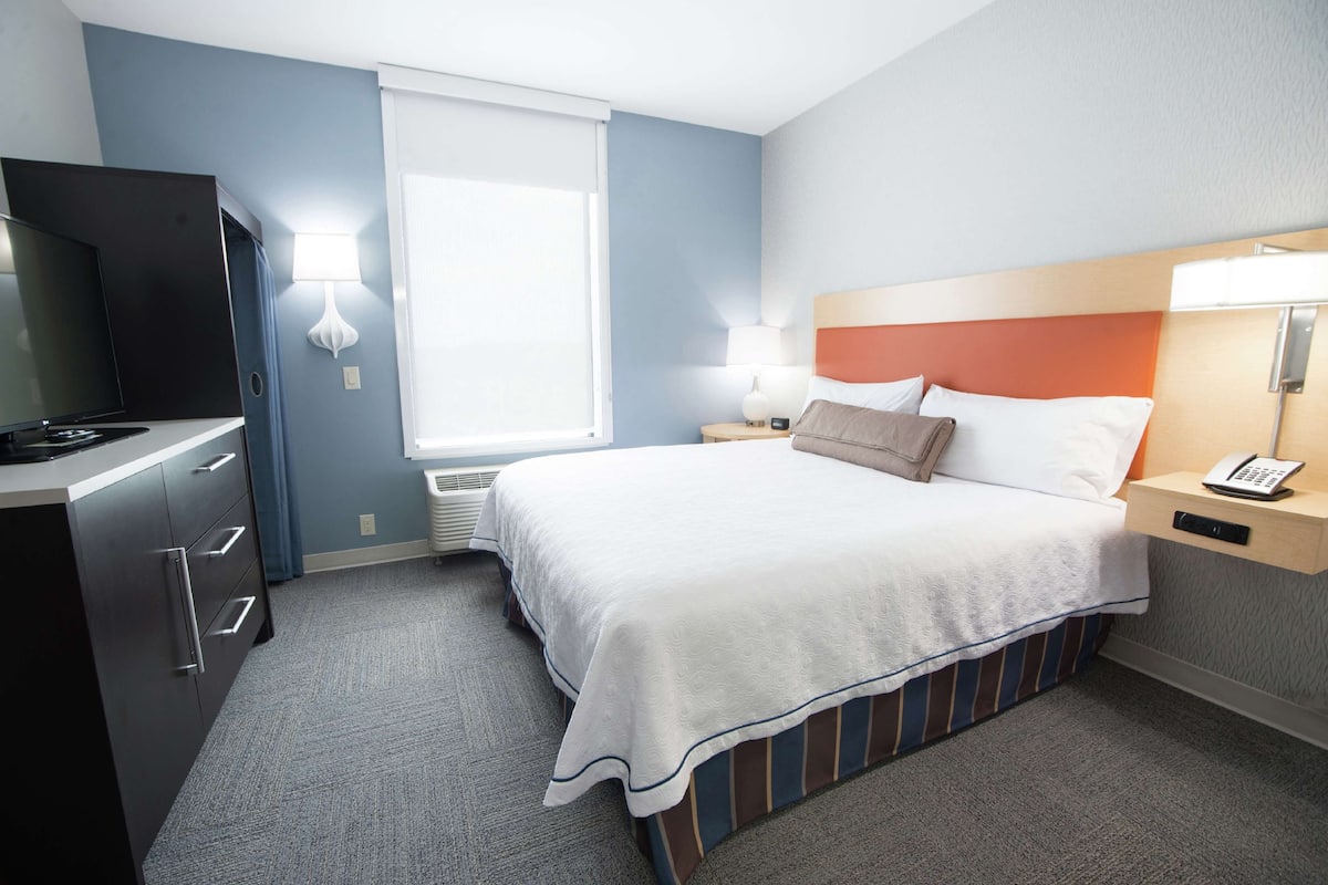 A hotel room with a bed, bedside table, lamp, TV, and a desk with drawers.